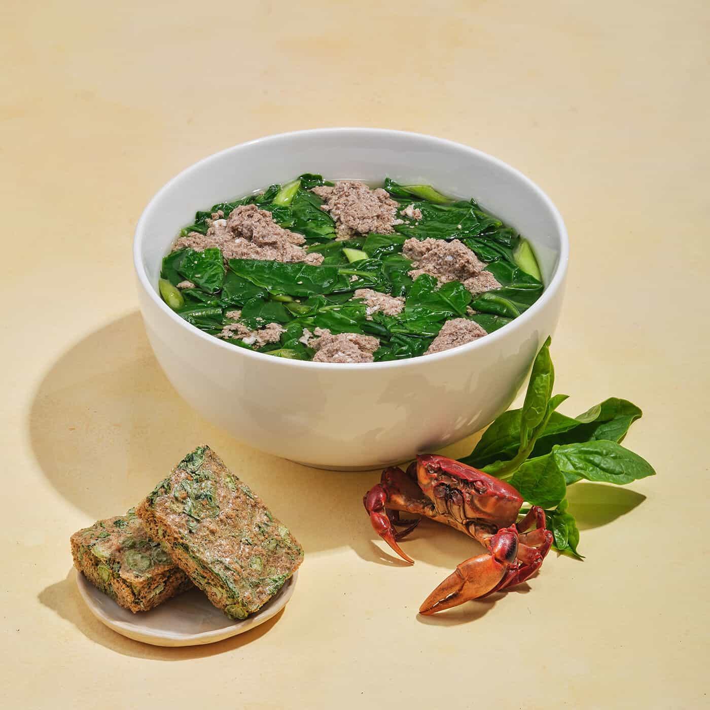 Bowl of crab meat soup with leafy greens, crab shell, fresh spinach, and spinach fritters on plate