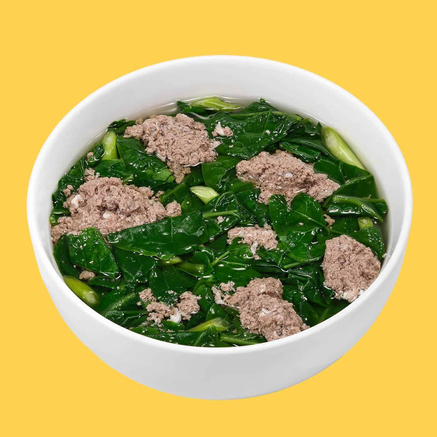 White bowl of clear broth soup with green leafy vegetables and chunks of minced crab meat on yellow background