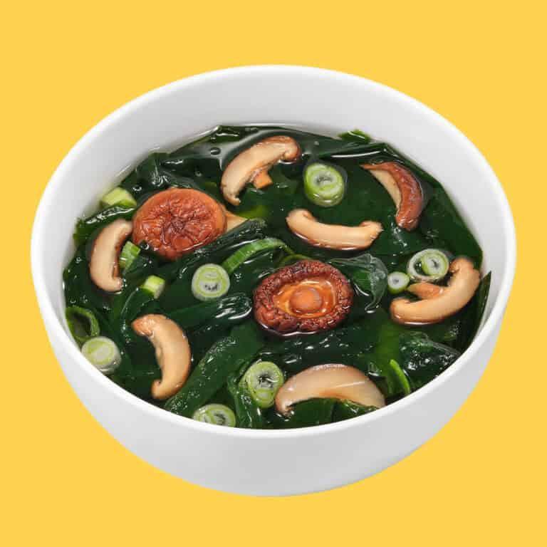 White bowl of miso soup with shiitake mushrooms, green onions, and seaweed on yellow background