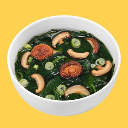 White bowl of miso soup with shiitake mushrooms, green onions, and seaweed on yellow background