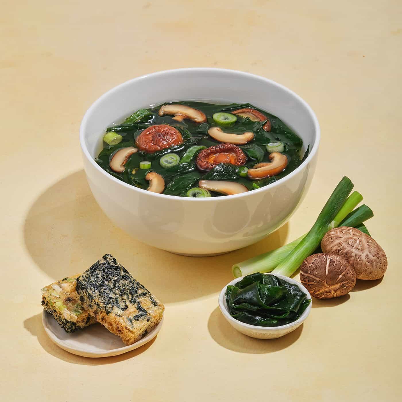 White bowl of miso soup with shiitake mushrooms, seaweed, green onions, and vegetable tempura on beige surface