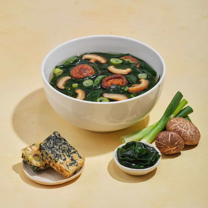 White bowl of miso soup with shiitake mushrooms, seaweed, green onions, and vegetable tempura on beige surface